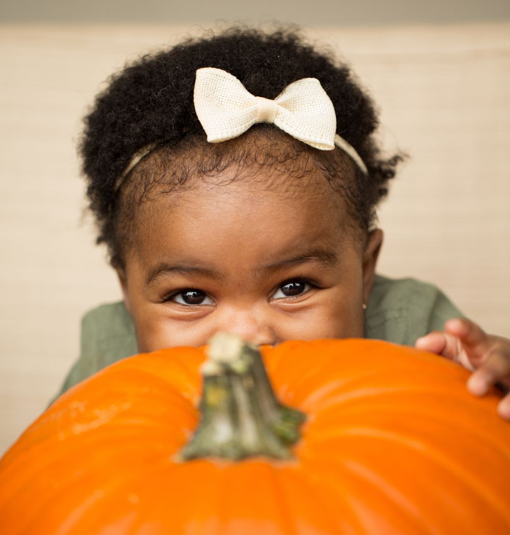Baby wearing a cream bow headband peeking over a large orange pumpkin